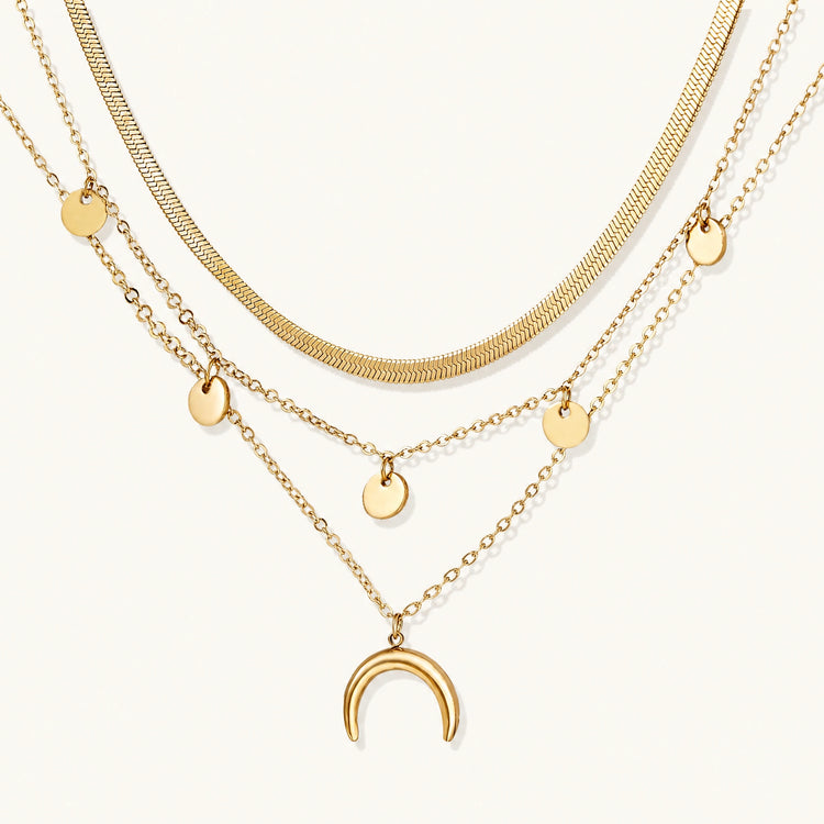 Four-layer gold necklace with herringbone chain, disc charms, and crescent moon pendant on cream background