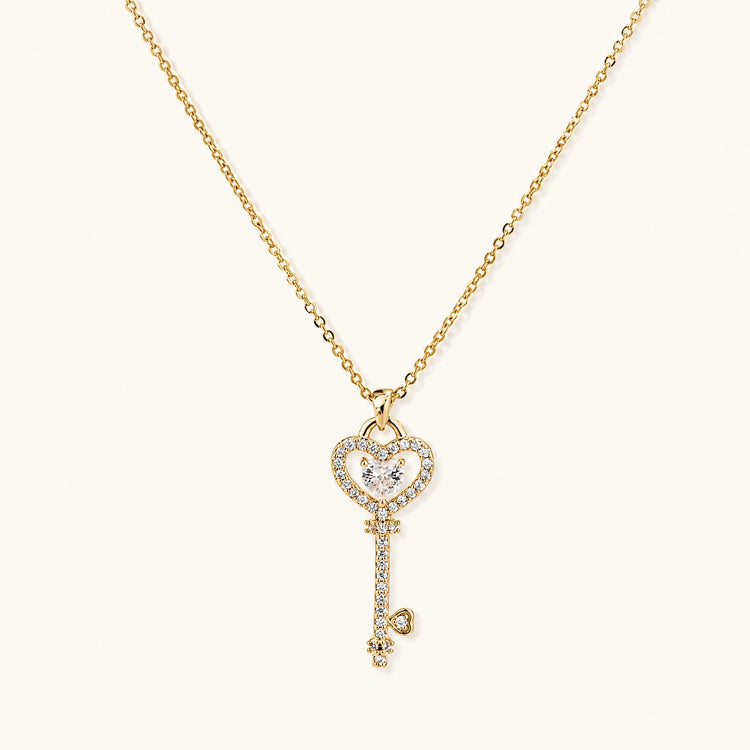 Delicate gold key pendant with heart-shaped bow and crystal pavé detailing on fine chain against cream background.