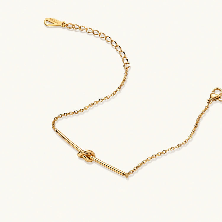 Delicate gold chain bracelet with minimalist bar and centered knot detail on cream background